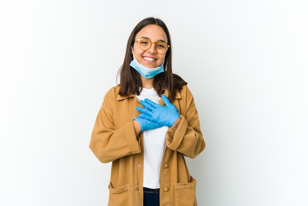 Jovem mulher latina usando uma máscara para se proteger de cobiçoso isolado no fundo branco tem uma expressão amigável, pressionando a palma da mão no peito. Conceito de amor.