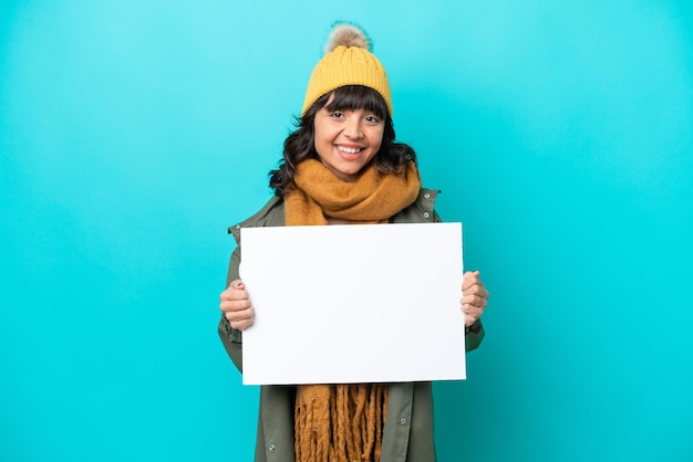 Jovem mulher latina vestindo jaqueta de inverno isolada em fundo azul segurando um cartaz vazio com expressão feliz