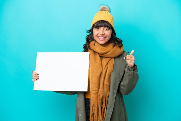 Jovem mulher latina vestindo jaqueta de inverno isolada em fundo azul segurando um cartaz vazio com o polegar para cima