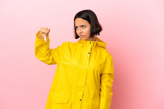 Jovem mulher latina vestindo um casaco à prova de chuva sobre um fundo isolado, mostrando o polegar para baixo com expressão negativa