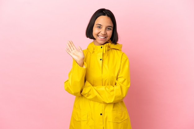 Jovem mulher latina vestindo um casaco à prova de chuva sobre um fundo isolado saudando com a mão com expressão feliz