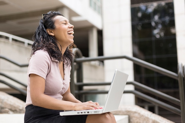Jovem mulher latino-americana muito sorridente ao ar livre. Ela está sentada usando um laptop.