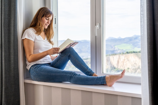 Jovem mulher lendo sentado na janela de sua casa. Conceito de trabalho ou estudo em casa.