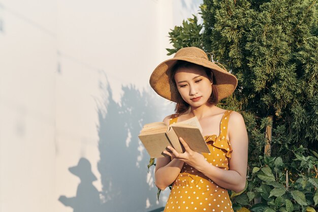 Jovem mulher lendo um livro em pé magro contra o tronco de uma árvore no parque de verão ao ar livre.