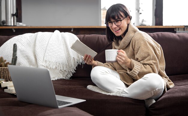 Jovem mulher linda com um suéter aconchegante com uma xícara e um livro nas mãos e um laptop no sofá em casa. conceitos de aprendizagem online.