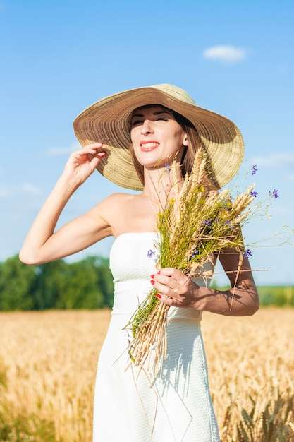 Jovem mulher linda em um vestido branco e um chapéu detém um buquê com flores silvestres em um campo de milho.