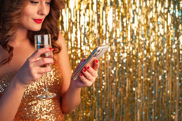 Jovem mulher linda em um vestido dourado bebe champanhe, segurando um smartphone, se divertindo em uma festa em um fundo dourado brilhante