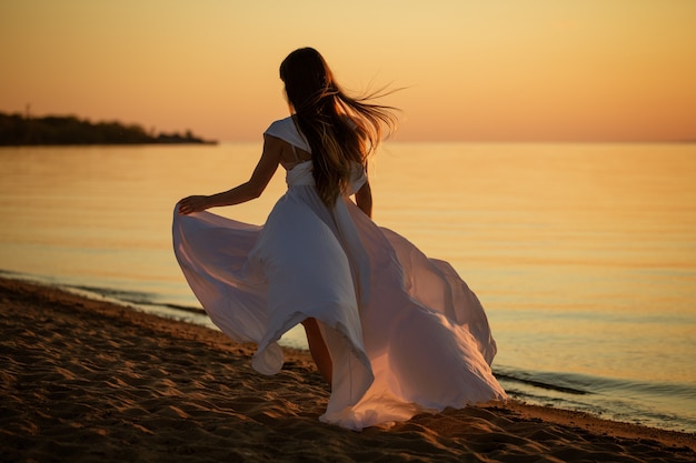 Jovem mulher livre correndo em um vestido branco perto do mar ao pôr do sol