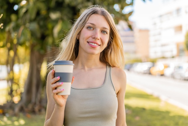 Jovem mulher loira ao ar livre segurando um café para viagem com expressão feliz