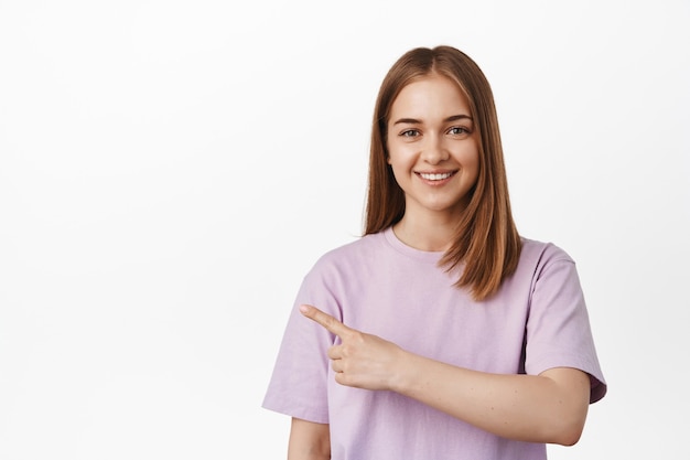 Jovem mulher loira apontando o dedo para a esquerda, sorrindo feliz, em pé com uma camiseta contra uma parede branca