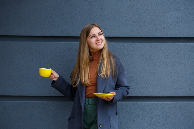 Jovem mulher loira bonita com um casaco cinza bebe chá quente em uma xícara amarela em um dia frio de outono. deliciosa bebida quente na esplanada do café