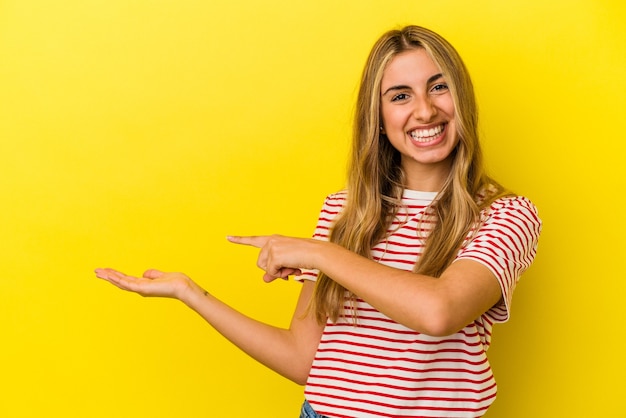 Jovem mulher loira caucasiana isolada em fundo amarelo animado segurando um espaço de cópia na palma da mão.