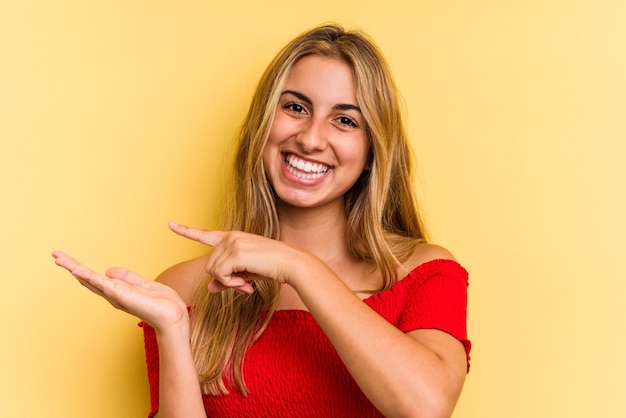 Jovem mulher loira caucasiana isolada em fundo amarelo animado segurando um espaço de cópia na palma da mão.