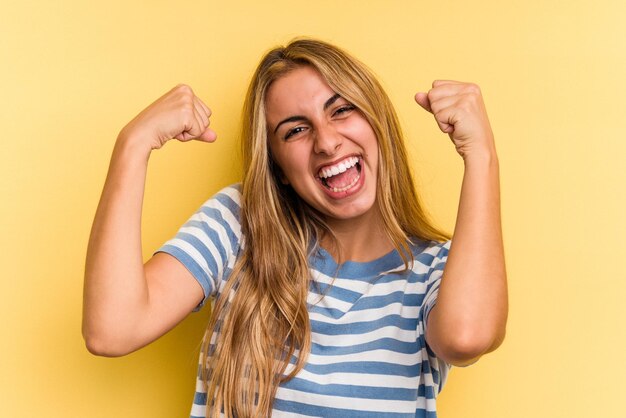 Jovem mulher loira caucasiana isolada em fundo amarelo mostrando força gesto com os braços, símbolo do poder feminino