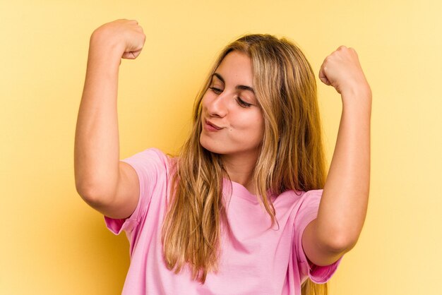 Jovem mulher loira caucasiana isolada em fundo amarelo mostrando força gesto com os braços, símbolo do poder feminino