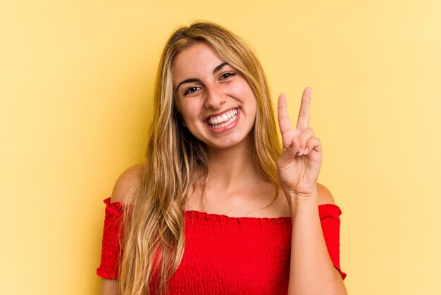 Jovem mulher loira caucasiana isolada em fundo amarelo, mostrando sinal de vitória e sorrindo amplamente.