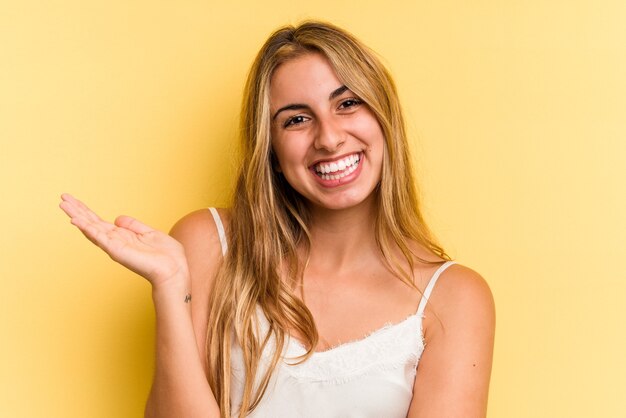 Jovem mulher loira caucasiana isolada em fundo amarelo, mostrando um espaço de cópia na palma da mão e segurando a outra mão na cintura.