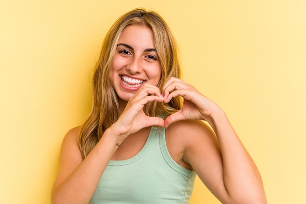 Jovem mulher loira caucasiana isolada em fundo amarelo, sorrindo e mostrando uma forma de coração com as mãos.