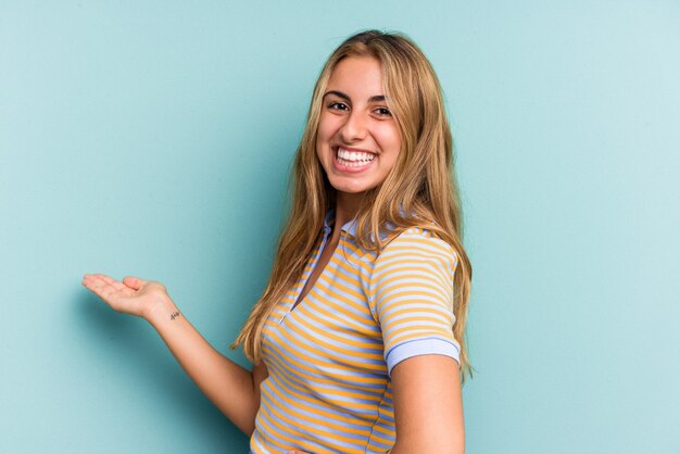 Jovem mulher loira caucasiana isolada em um fundo azul, mostrando uma expressão de boas-vindas.
