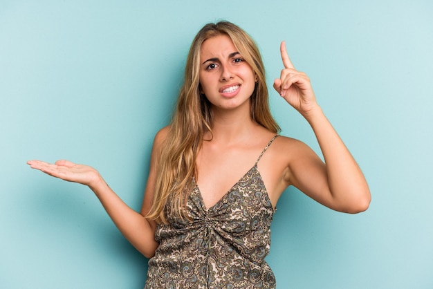 Jovem mulher loira caucasiana isolada em um fundo azul, segurando e mostrando um produto por lado.