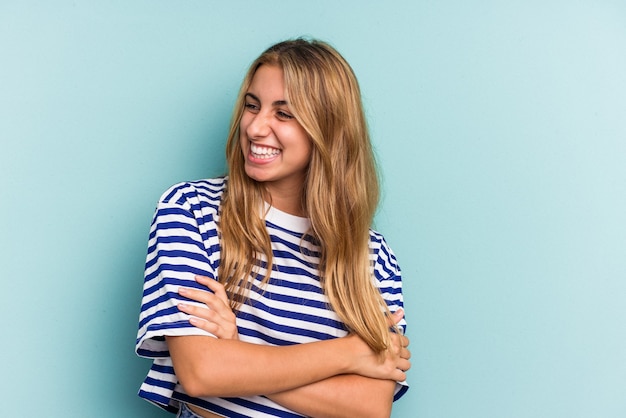 Jovem mulher loira caucasiana isolada em um fundo azul, sorrindo confiante com os braços cruzados.