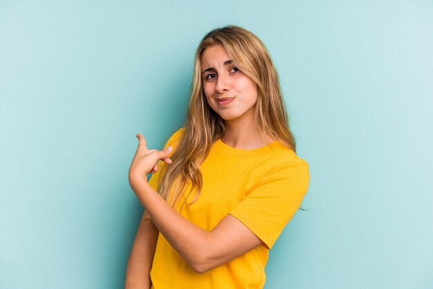 Jovem mulher loira caucasiana isolada em um fundo azul, sorrindo e apontando de lado, mostrando algo no espaço em branco.