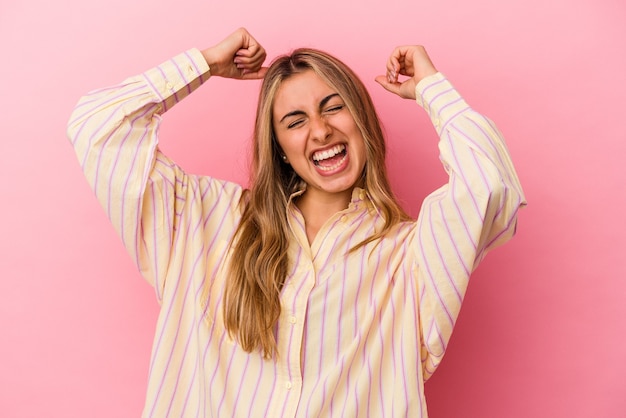 Jovem mulher loira caucasiana isolada em um fundo rosa, comemorando um dia especial, pula e levanta os braços com energia.