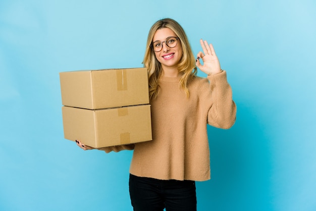 Jovem mulher loira caucasiana segurando caixas para mover-se alegre e confiante, mostrando um gesto de ok.