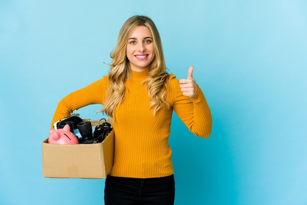 Jovem mulher loira caucasiana segurando caixas para se mover sorrindo e levantando o polegar