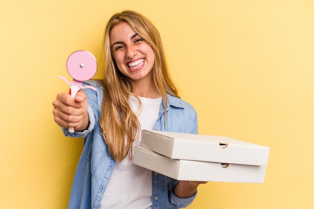 Jovem mulher loira caucasiana segurando pizzas e cortador isolado em fundo amarelo