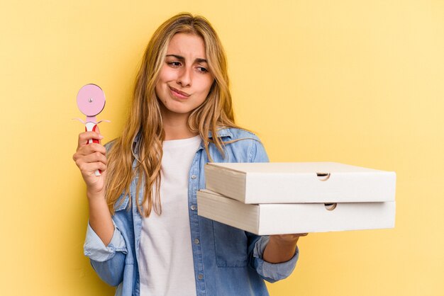 Jovem mulher loira caucasiana segurando pizzas e cortador isolados em fundo amarelo