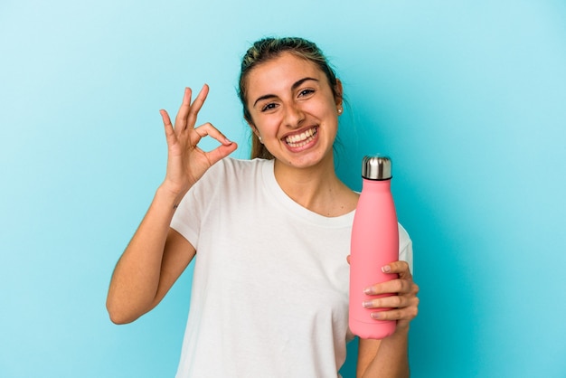 Jovem mulher loira caucasiana segurando uma garrafa térmica isolada sobre fundo azul alegre e confiante mostrando um gesto ok.