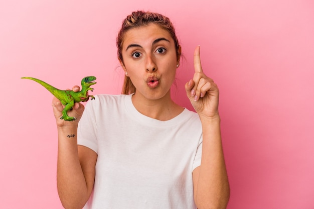 Jovem mulher loira caucasiana segurando uma miniatura de dinossauro isolada na parede rosa, tendo uma ótima ideia, o conceito de criatividade.