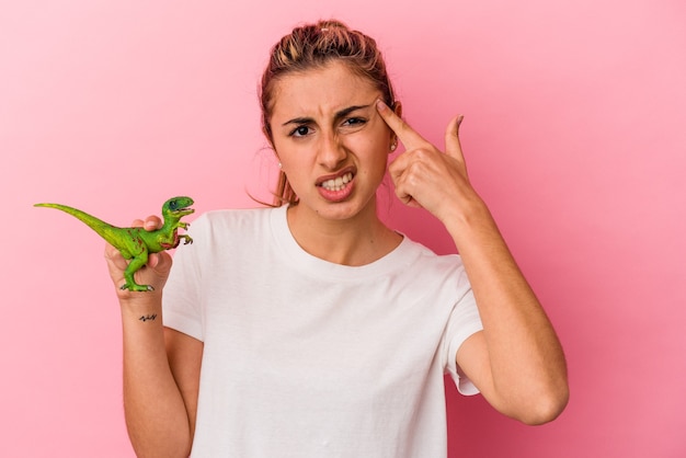 Jovem mulher loira caucasiana segurando uma miniatura de dinossauro isolada no fundo rosa, mostrando um gesto de decepção com o dedo indicador.