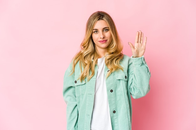 Jovem mulher loira caucasiana, sorrindo alegre mostrando o número cinco com os dedos.