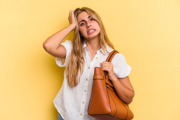 Jovem mulher loira caucasiana, vestindo uma bolsa de couro isolada em um fundo amarelo, sendo chocada, ela se lembrou de uma reunião importante.