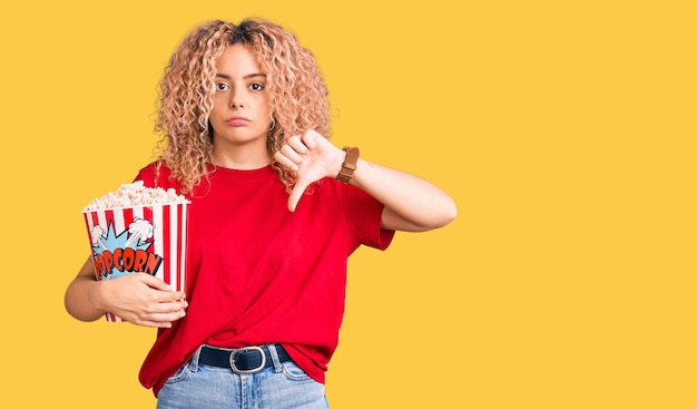 Jovem mulher loira com cabelo encaracolado comendo pipoca com sinal negativo de rosto zangado, mostrando desagrado com o conceito de rejeição de polegares para baixo