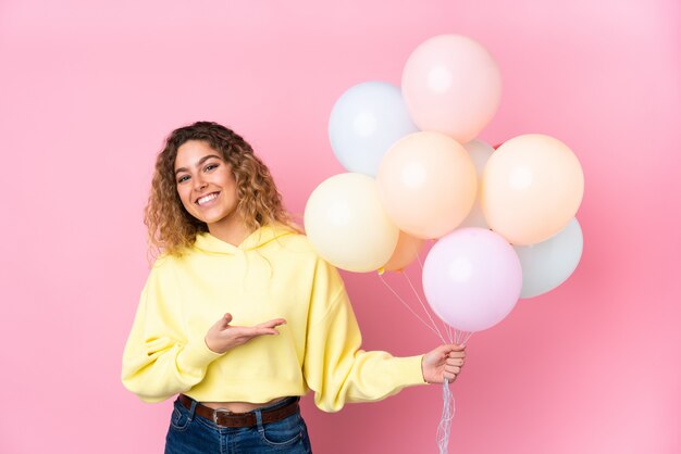 Jovem mulher loira com cabelos cacheados, pegando muitos balões na parede rosa, estendendo as mãos para o lado