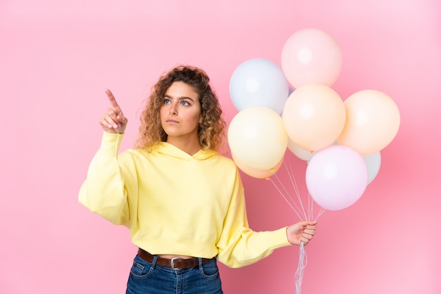 Jovem mulher loira com cabelos cacheados, pegando muitos balões na parede rosa tocando na tela transparente
