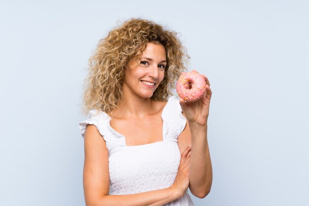 Jovem mulher loira com cabelos cacheados, segurando um donut sobre parede azul, olhando para cima enquanto sorrindo