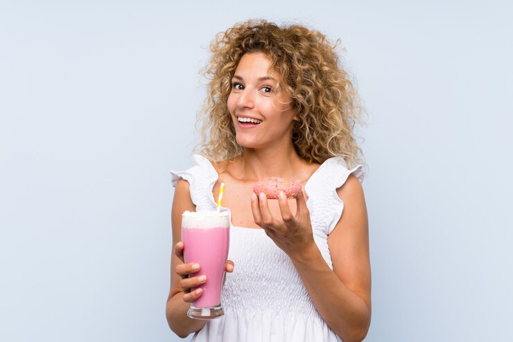 Foto jovem mulher loira com cabelos cacheados, segurando um milk-shake de morango e um donut