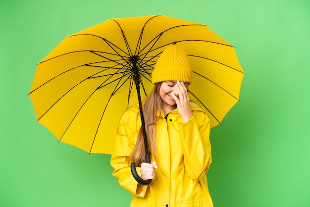 Jovem mulher loira com casaco à prova de chuva e guarda-chuva sobre fundo croma isolado rindo