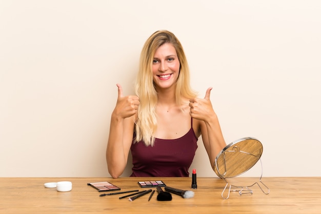 Jovem mulher loira com cosméticos em uma mesa dando um polegar para cima gesto