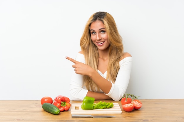 Jovem mulher loira com legumes em uma mesa, apontando o dedo para o lado