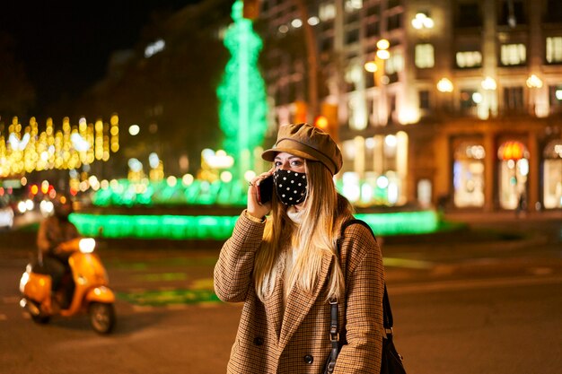 Jovem mulher loira com máscara falando ao telefone em uma cidade à noite. atmosfera de inverno.