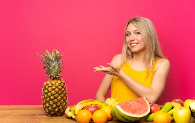 Jovem mulher loira com muitas frutas, estendendo as mãos para o lado para convidar para vir