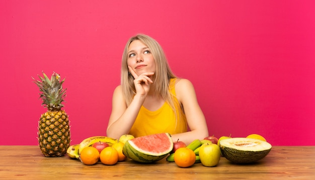 Jovem mulher loira com muitas frutas pensando uma idéia