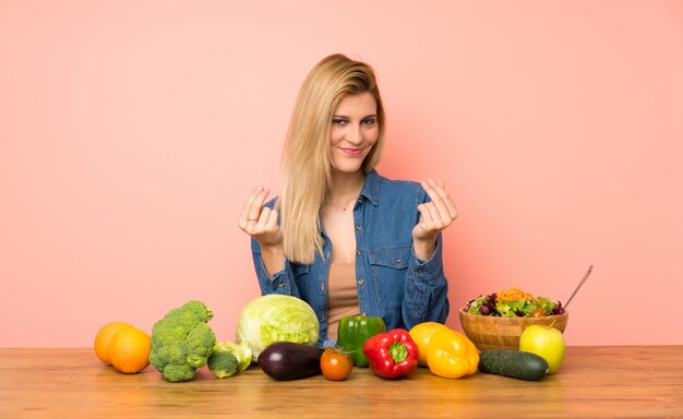 Jovem mulher loira com muitos legumes fazendo dinheiro gesto