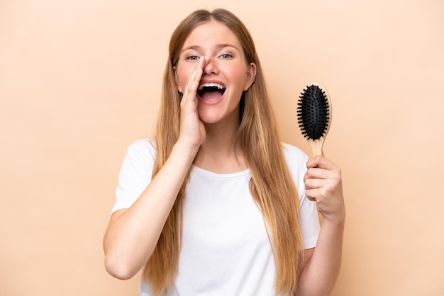 Jovem mulher loira com pente de cabelo isolado em fundo bege gritando com a boca aberta