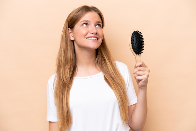 Jovem mulher loira com pente de cabelo isolado em fundo bege olhando para cima enquanto sorria